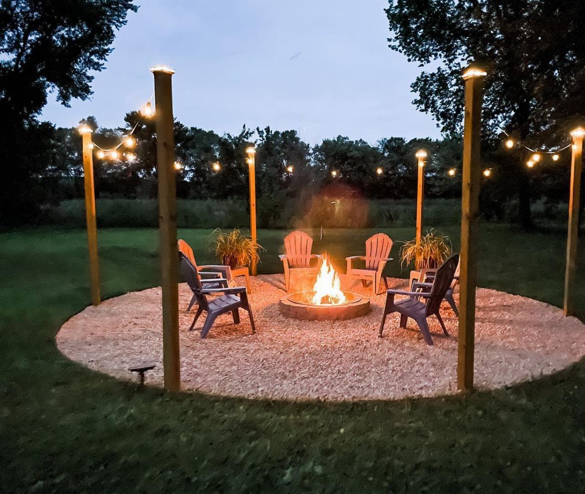A glowing fire pit surrounded by chairs under hanging string lights in a backyard.