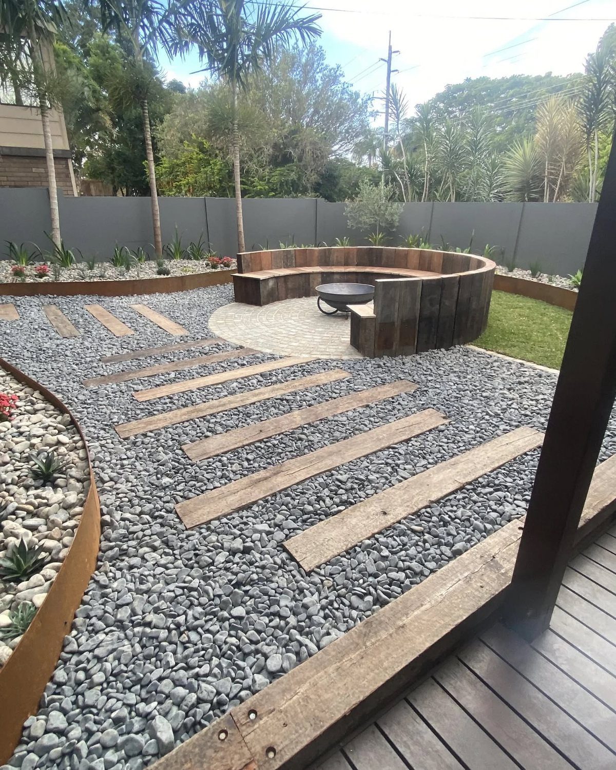 A semi-circle wooden bench surrounds a fire pit in a gravel backyard landscape.