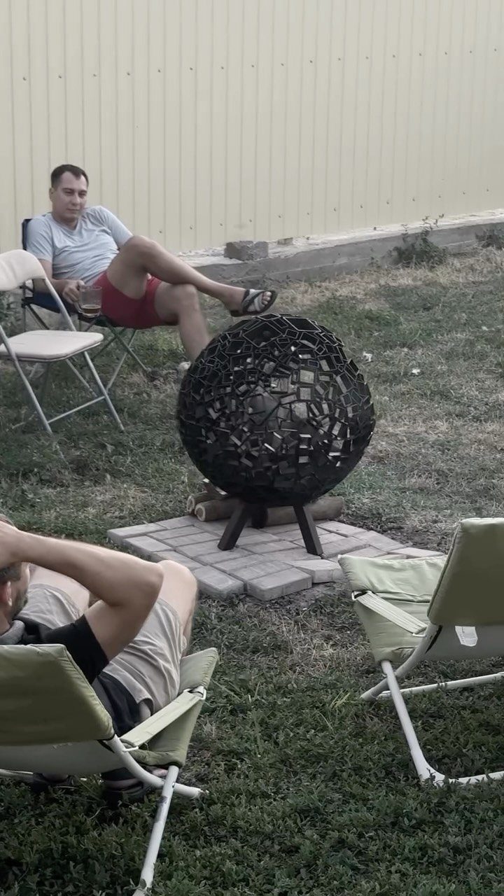 A black spherical metal fire pit sits on a tripod over stone pavers in a yard.