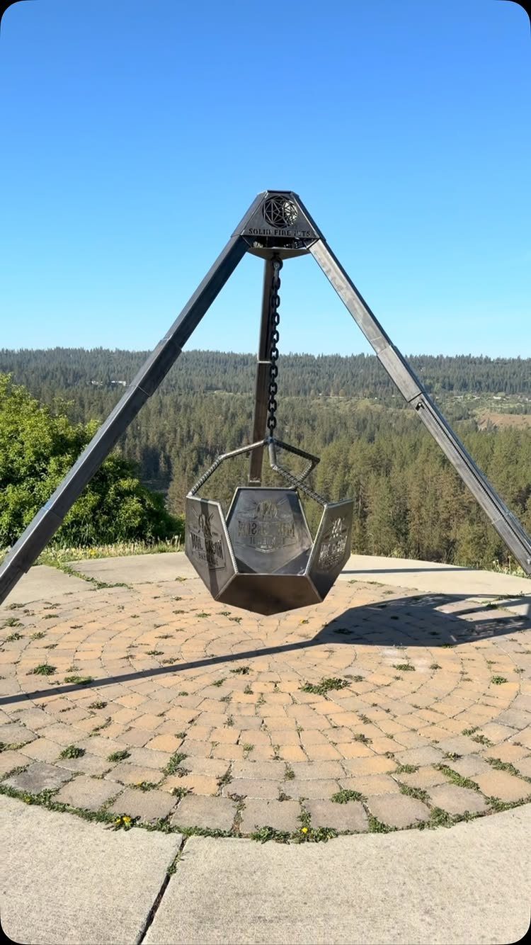 Metal tripod stand with a hanging fire bowl suspended over a stone patio overlooking a forest.