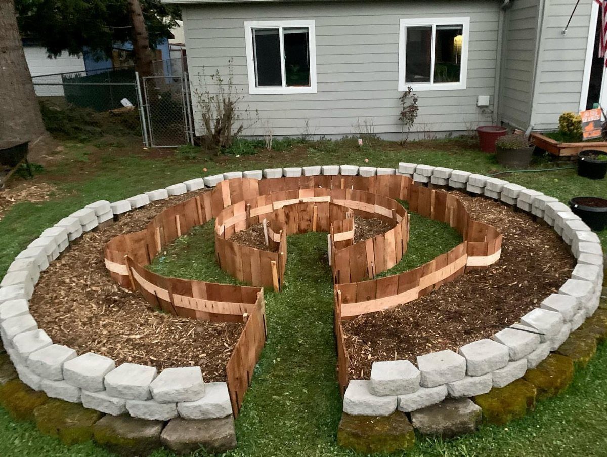 A circular garden bed with a stone border and wooden spiral dividers filled with dark mulch.