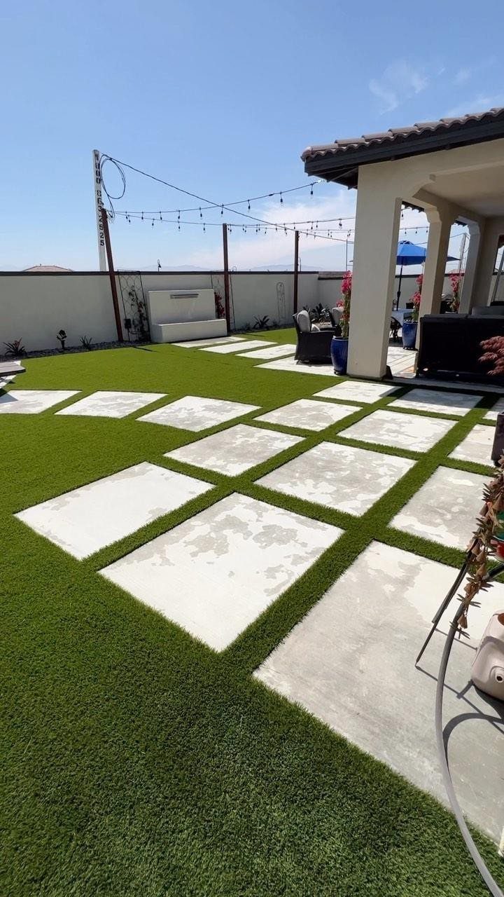 Large rectangular concrete stepping stones arranged in a grid pattern across green artificial turf in a backyard.