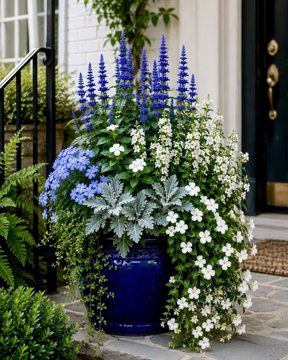 A vibrant blue ceramic planter filled with tall blue flowers, white blossoms, and silvery foliage on a porch.