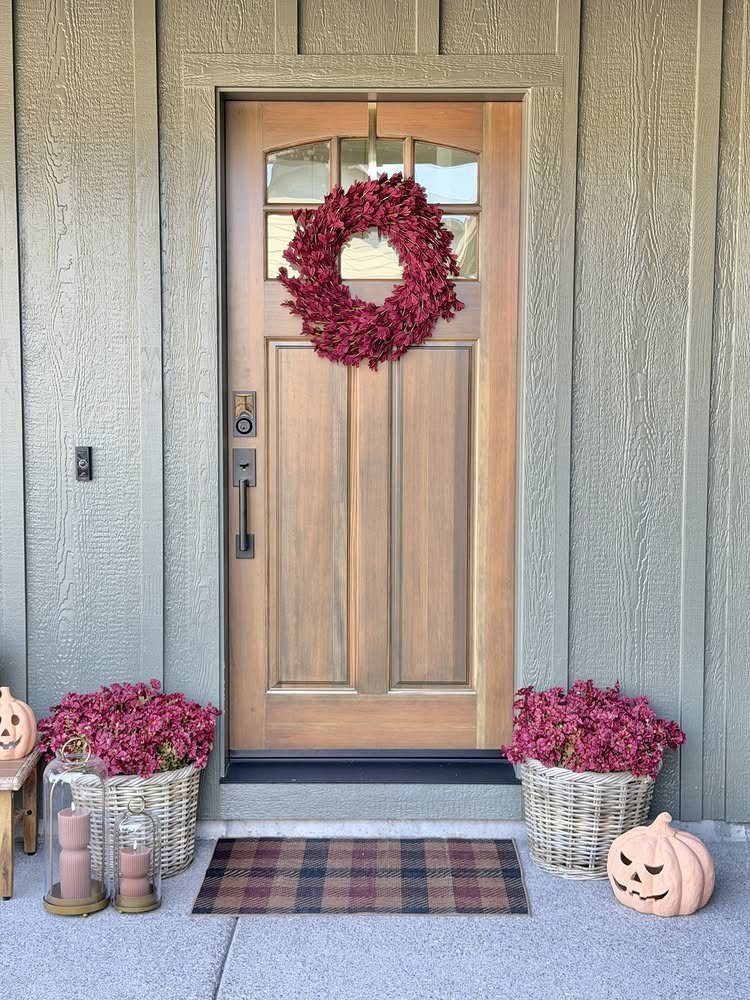A wooden front door decorated with a red wreath, flanked by flowering baskets and pumpkins.