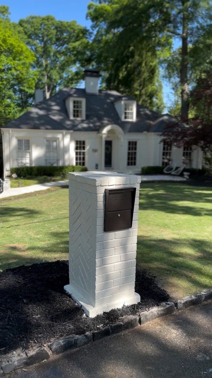 A white brick pedestal mailbox with a black metal slot sits in dark mulch.