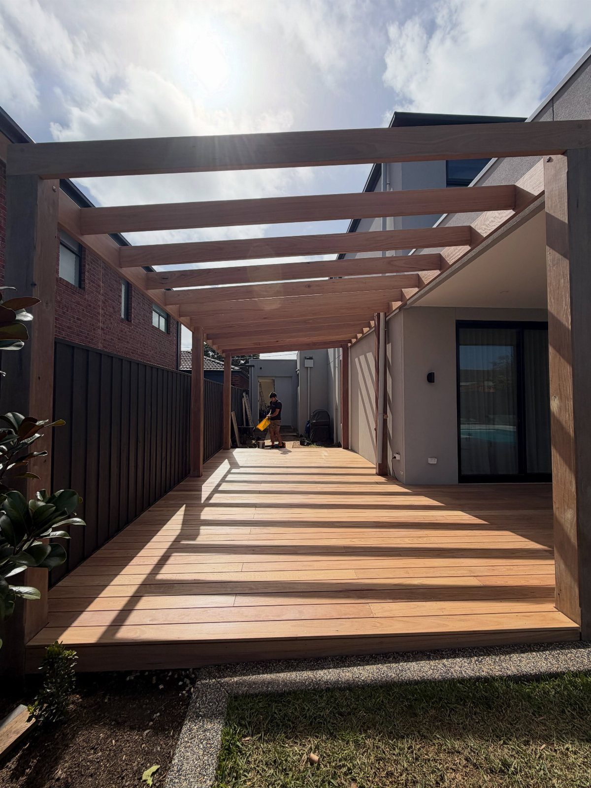A wooden pergola structure attached to a house covering a large timber deck in a backyard.