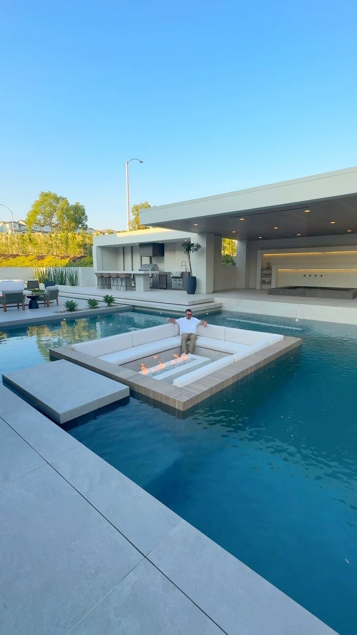 15 Decks and Porches Back Patio Ideas for Outdoor Living 5 A modern square firepit with white seating sits on a platform inside a large swimming pool.