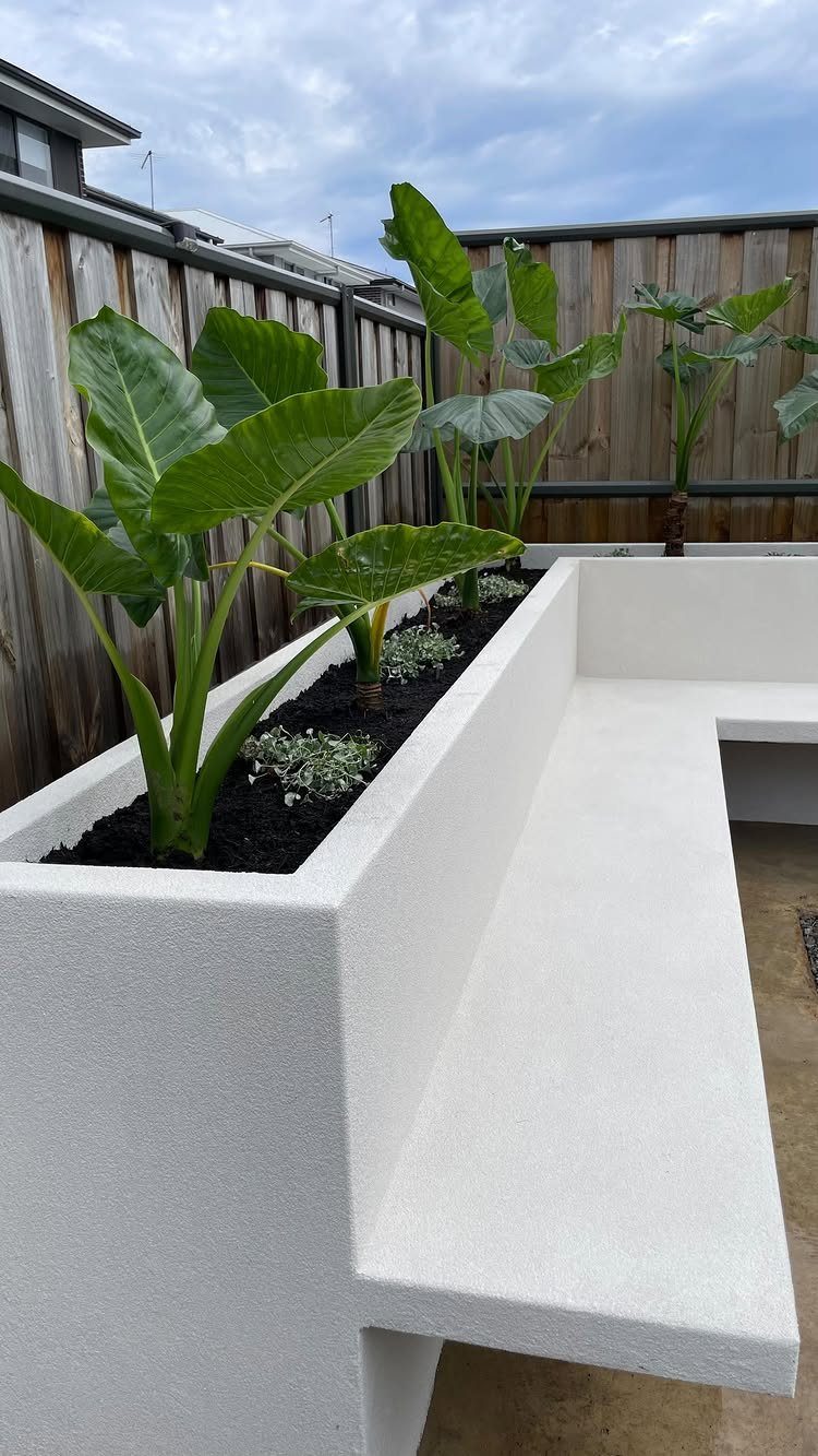 19 DIY Landscaping Ideas on a Budget for Any Yard 11 White concrete L-shaped bench with integrated planter containing large green elephant ear plants.