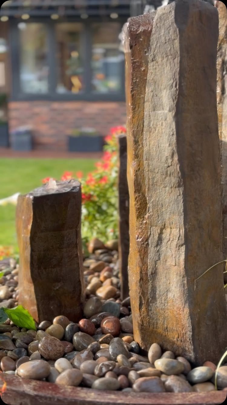 19 DIY Landscaping Ideas on a Budget for Any Yard 6 Vertical brown stones with water flowing over them surrounded by smooth river pebbles.