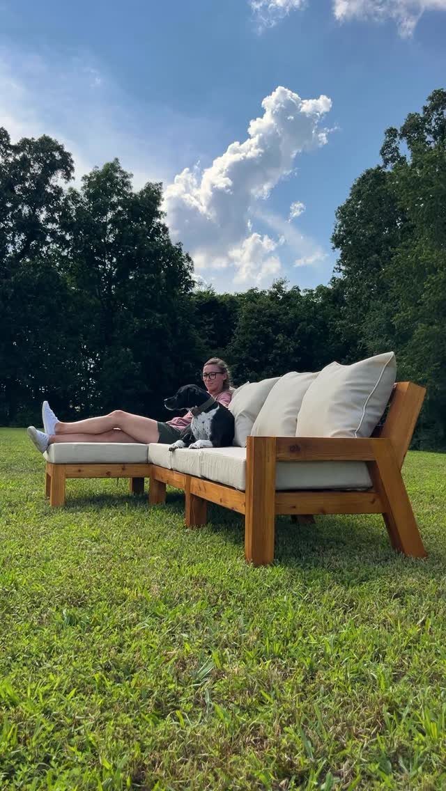 A wooden L-shaped outdoor sectional with cream cushions sits on a grassy lawn with a woman and dog.