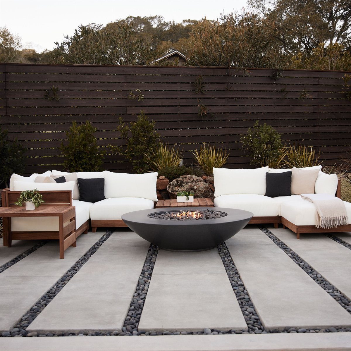 White cushioned outdoor sectional sofa with wooden frames surrounding a black fire pit on a patio.