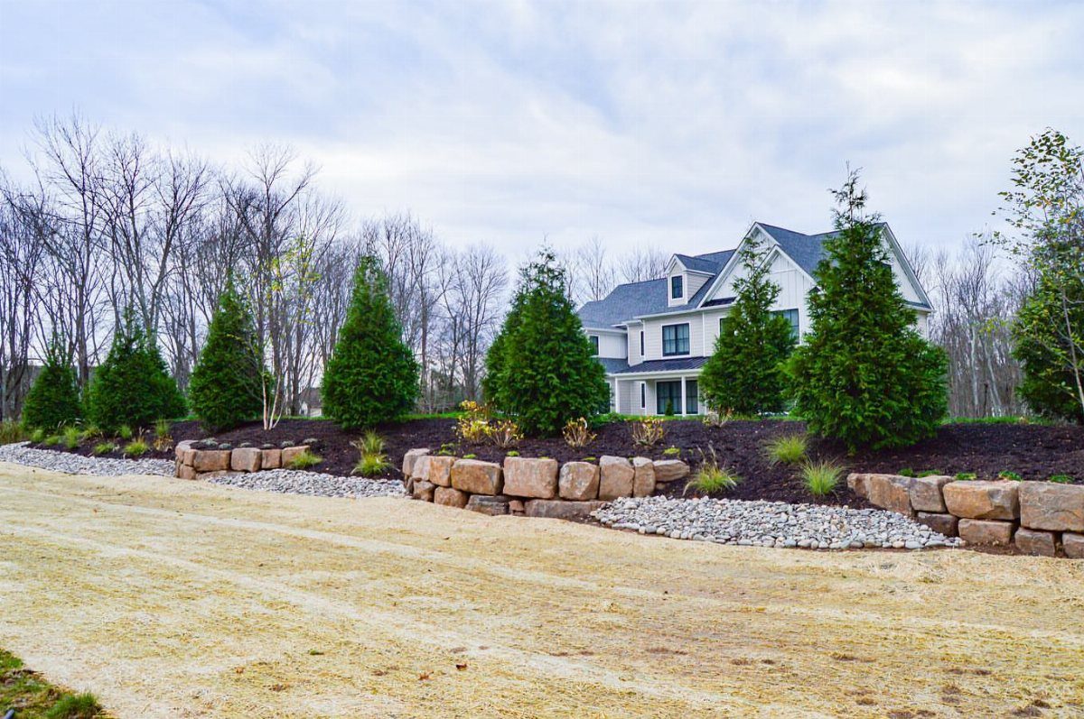 18 Farmhouse Backyard Ideas with Fire Pits, Herb Gardens & Outdoor Kitchens 2 Evergreen trees planted on a landscaped berm with stone retaining walls in front of a house.