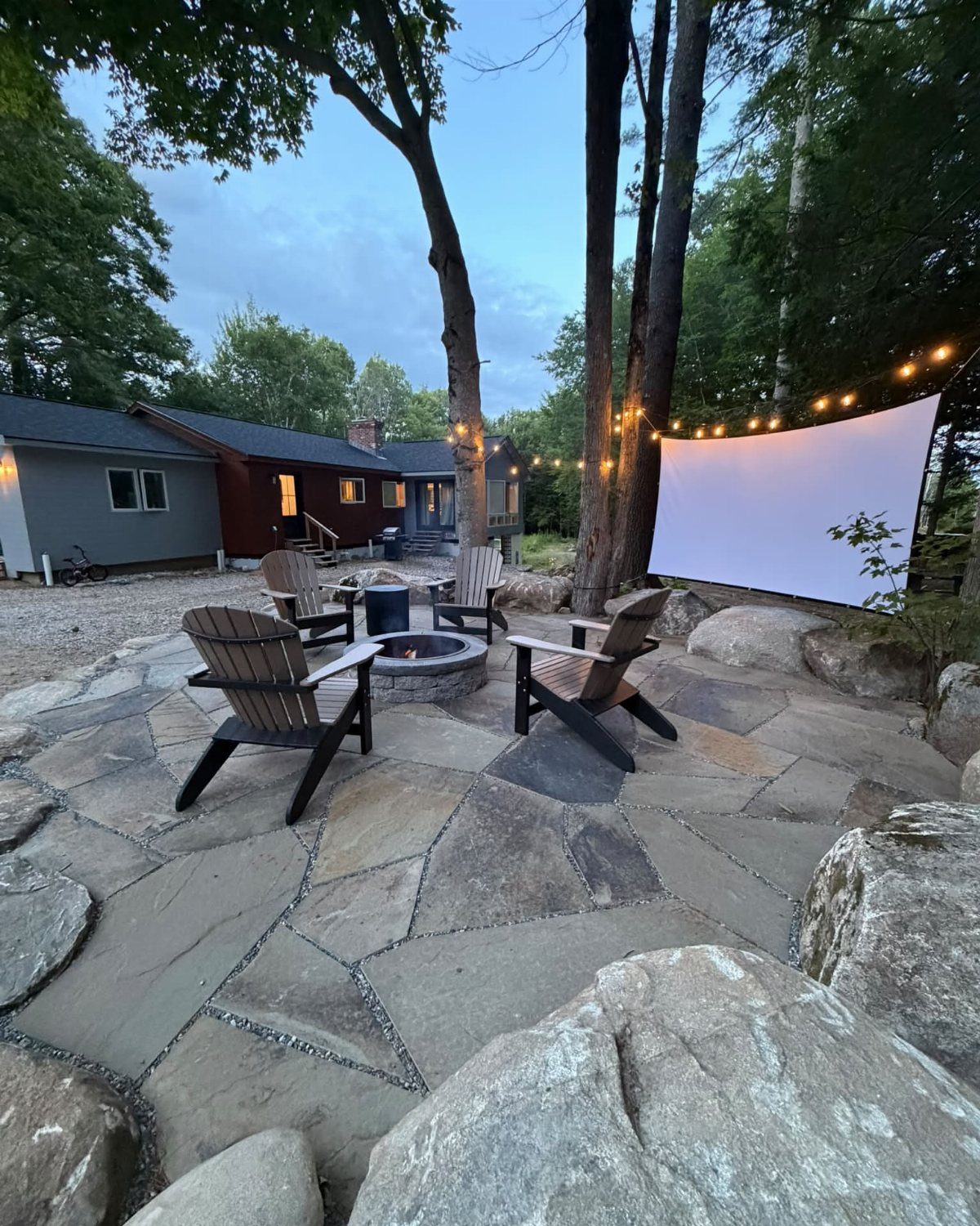 18 Farmhouse Backyard Ideas with Fire Pits, Herb Gardens & Outdoor Kitchens 3 Stone patio with a metal fire pit and four wooden Adirondack chairs under string lights.