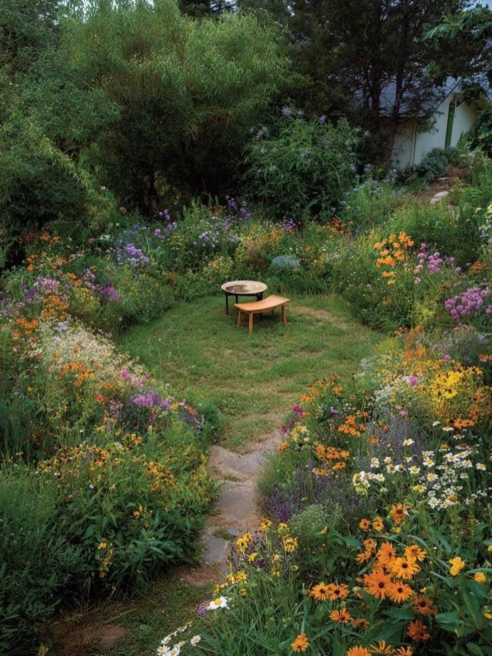 18 Farmhouse Backyard Ideas with Fire Pits, Herb Gardens & Outdoor Kitchens 17 Colorful wildflowers in orange, yellow, and purple surround a small wooden bench in a lush garden.