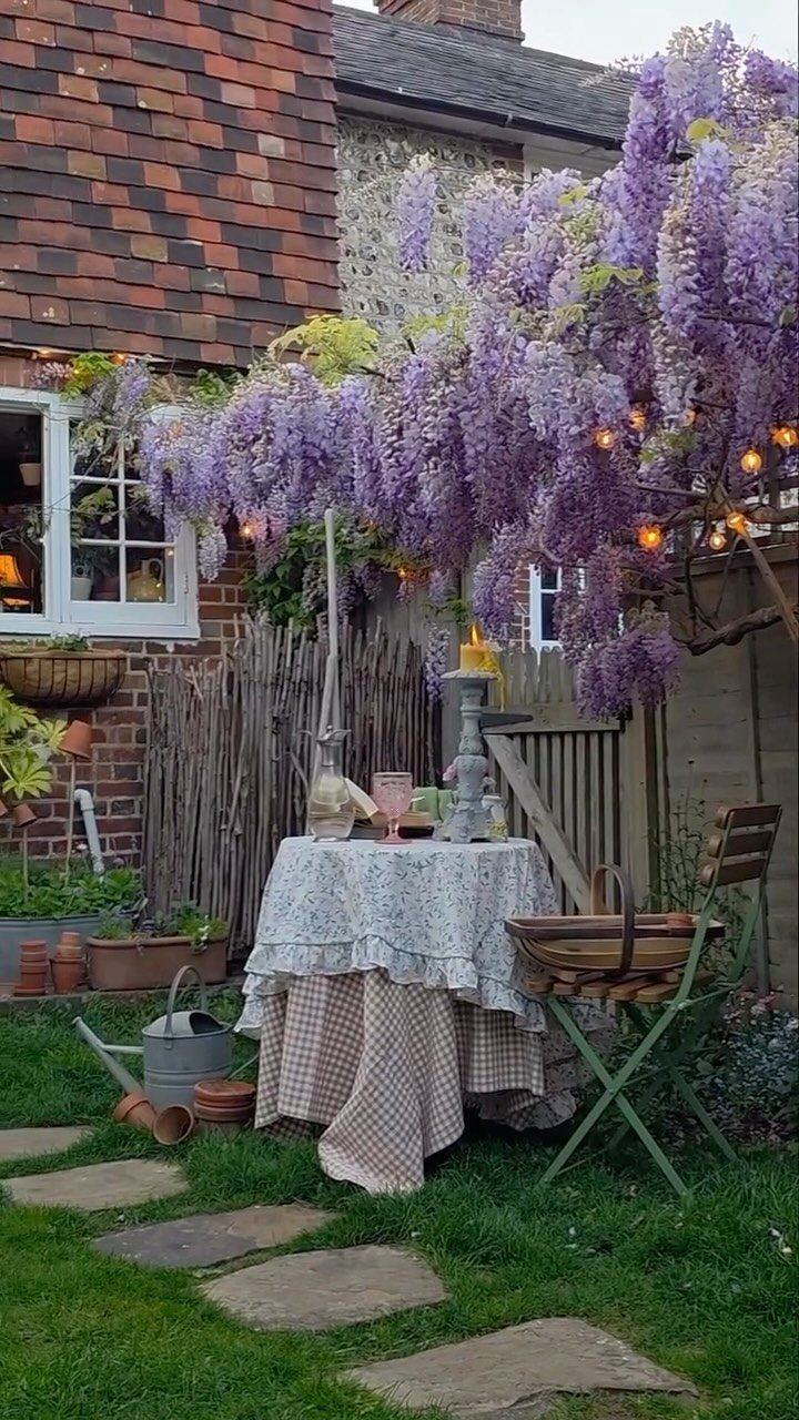 18 Farmhouse Backyard Ideas with Fire Pits, Herb Gardens & Outdoor Kitchens 18 A small table with a patterned cloth set under blooming purple wisteria in a garden.