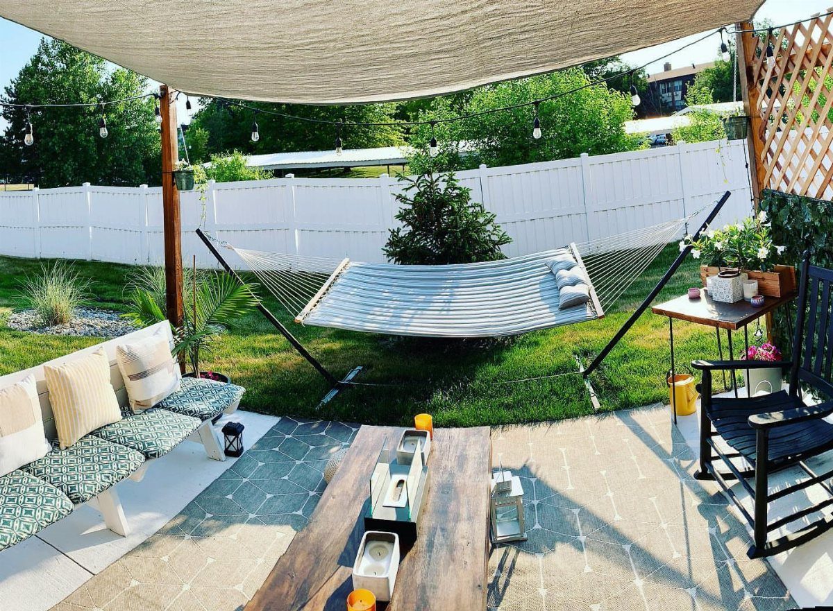 18 Farmhouse Backyard Ideas with Fire Pits, Herb Gardens & Outdoor Kitchens 13 A striped fabric hammock suspended between black metal poles in a lush green backyard.