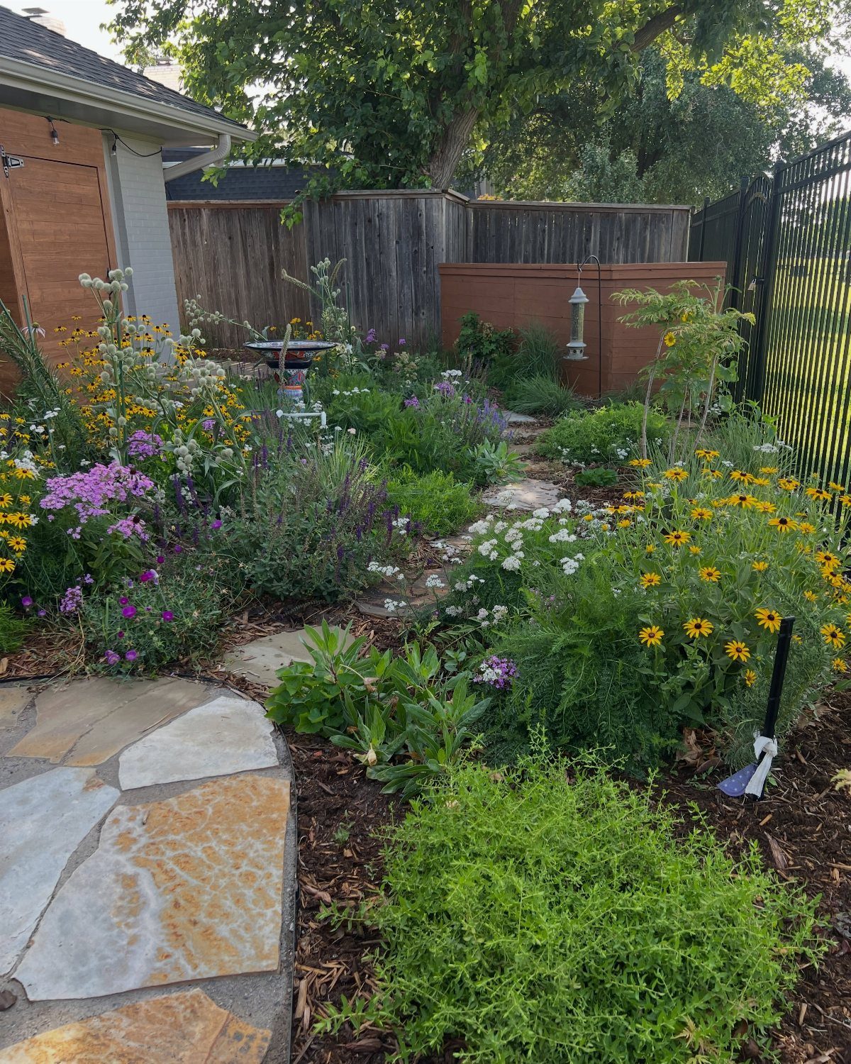 18 Farmhouse Backyard Ideas with Fire Pits, Herb Gardens & Outdoor Kitchens 12 Colorful wildflowers and green plants growing alongside a natural stone pathway in a backyard garden.