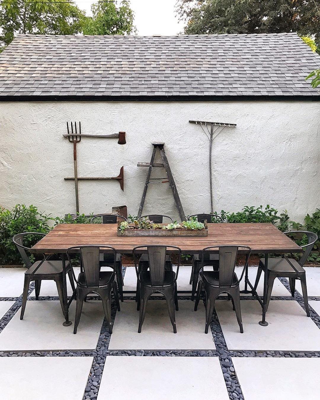 18 Farmhouse Backyard Ideas with Fire Pits, Herb Gardens & Outdoor Kitchens 16 A long wooden dining table with metal chairs set on a modern tiled patio outdoors.