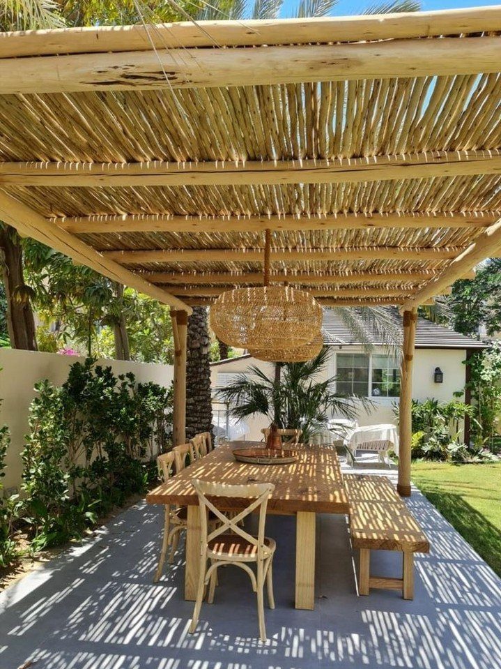 18 Farmhouse Backyard Ideas with Fire Pits, Herb Gardens & Outdoor Kitchens 14 Wooden dining set under a rustic pole pergola on a paved patio with greenery.