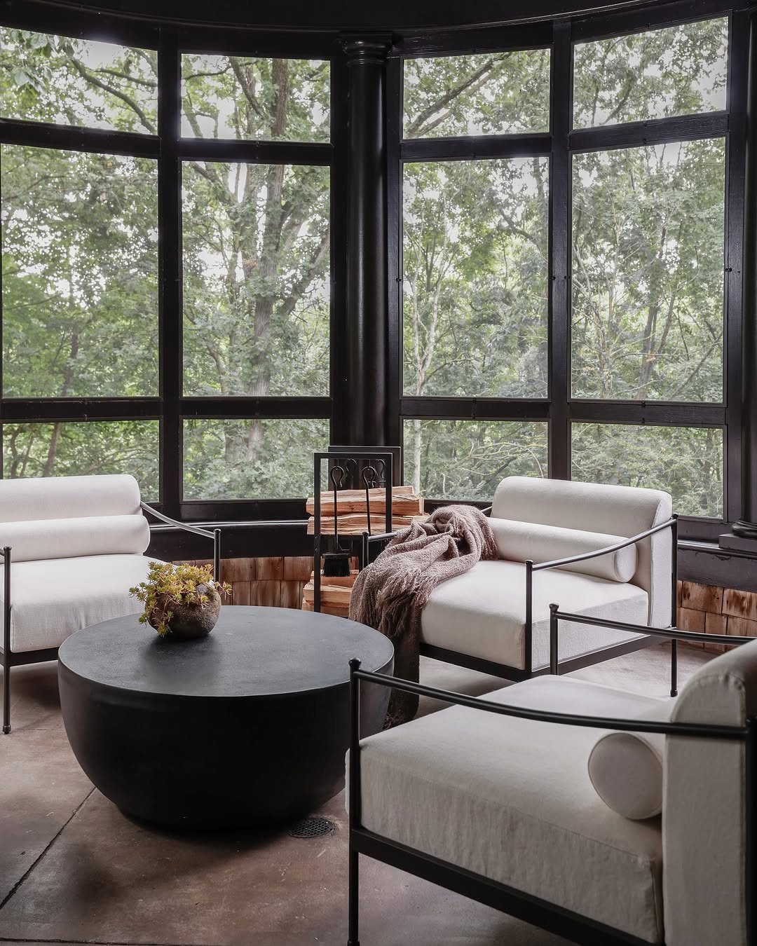 Modern sunroom with large windows, white armchairs, and a black coffee table overlooking green trees.