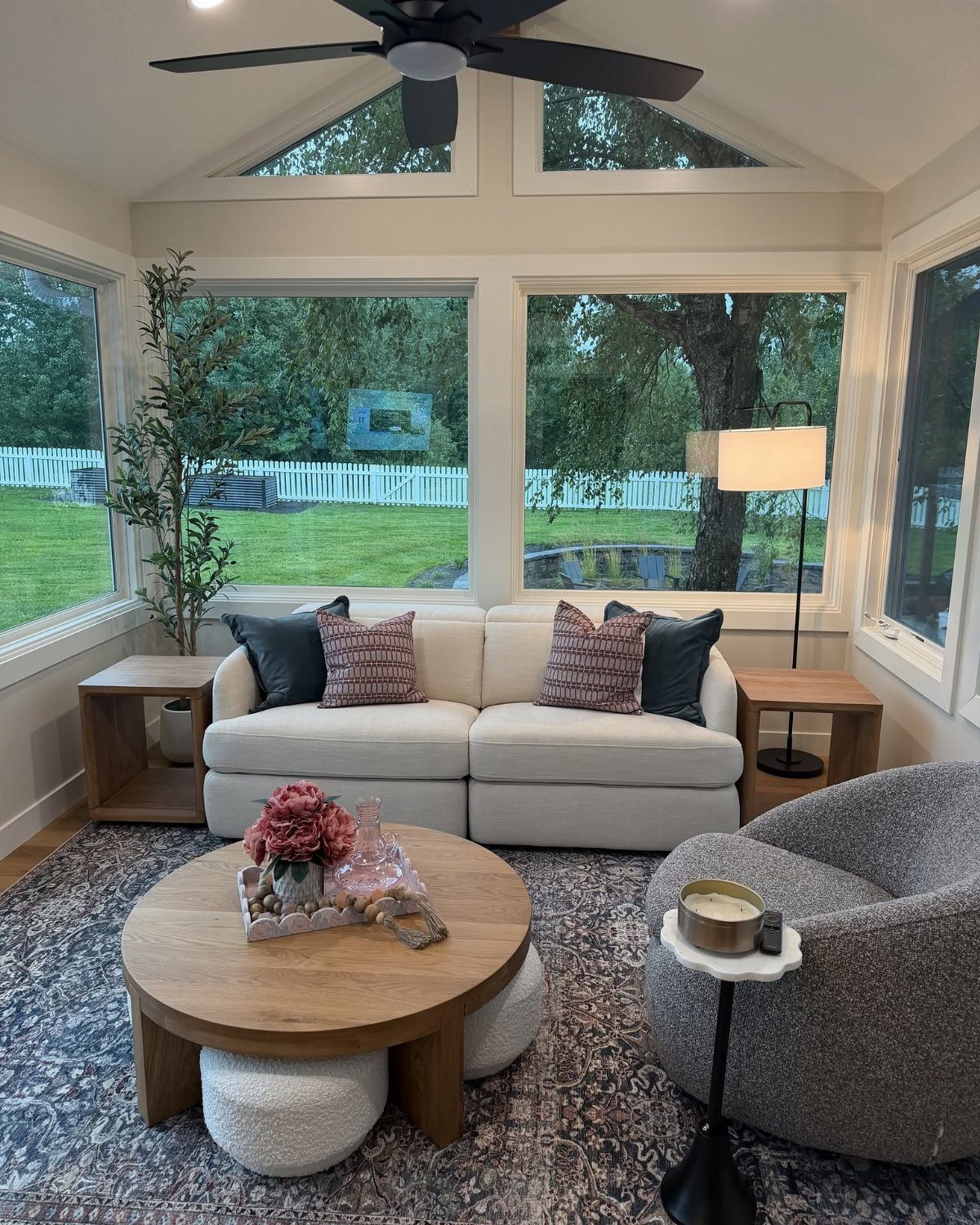 A cozy sunroom with white sofas, wooden tables, and large windows overlooking a green yard.