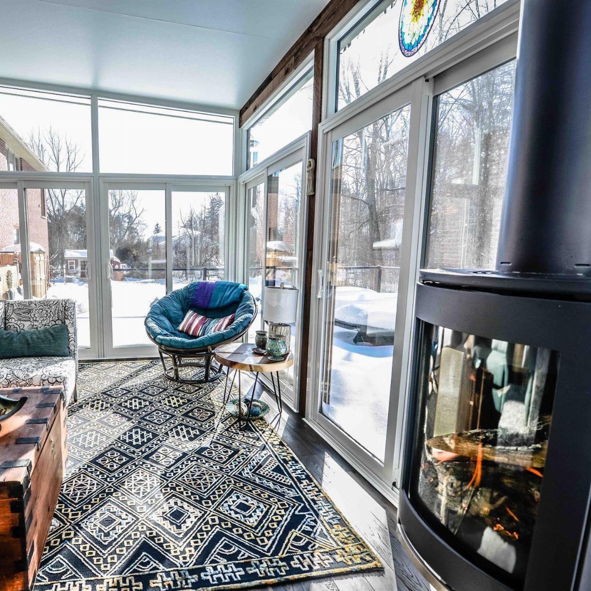 A cozy sunroom featuring a wood-burning stove, blue lounge chair, and patterned rug overlooking snow.