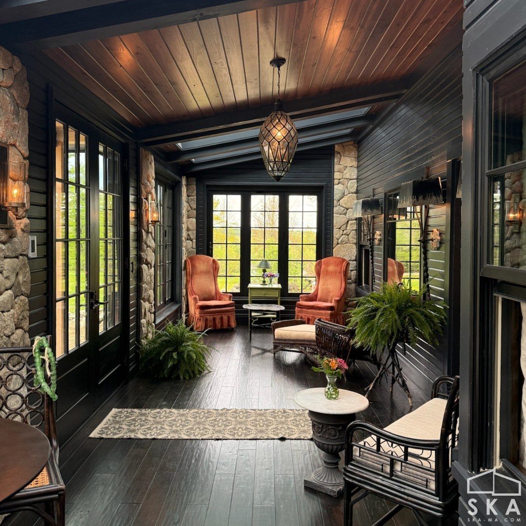A cozy sunroom with dark wood ceilings, stone walls, large windows, and comfortable orange armchairs.