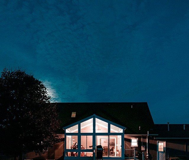 A glowing sunroom addition with large glass windows attached to a dark house at twilight.