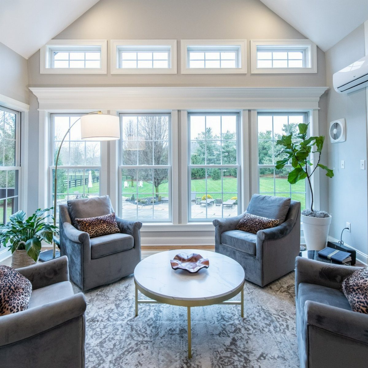 A bright sunroom with large windows, grey armchairs, and a marble coffee table.