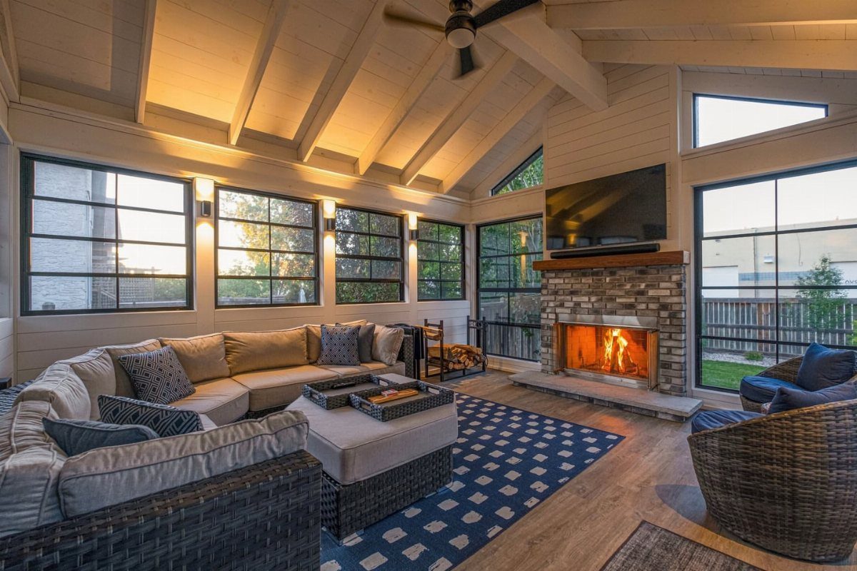 A cozy sunroom featuring large windows, a stone fireplace with a fire, and comfortable lounge seating.