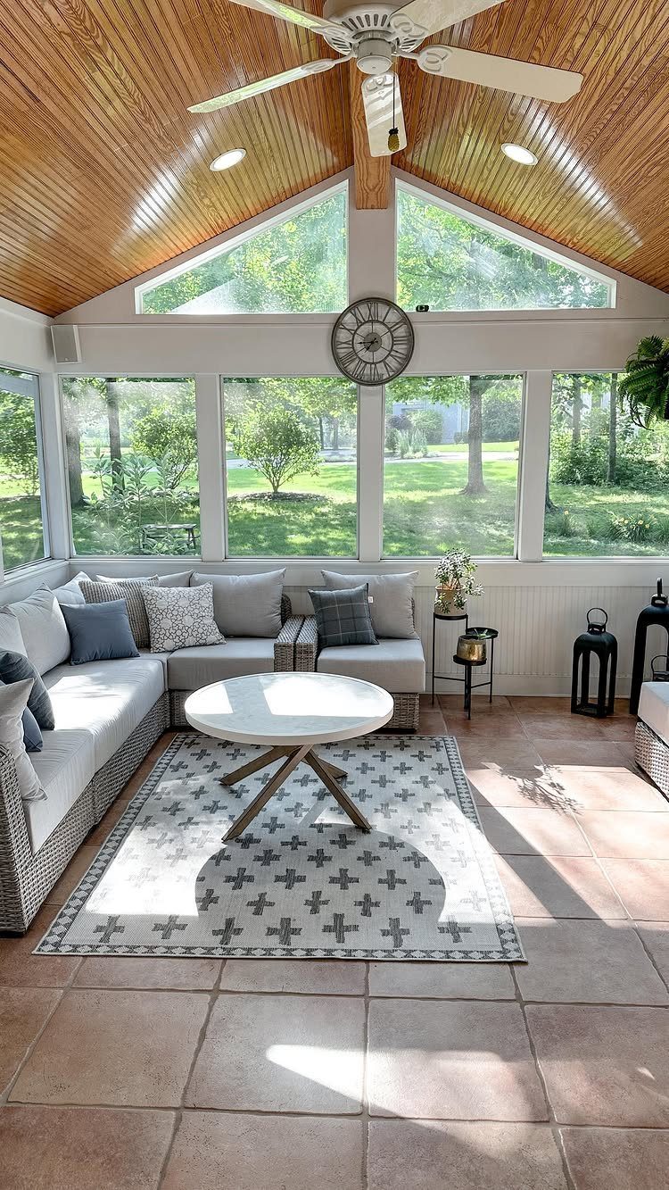 A bright sunroom with a wood ceiling, large windows, a grey sectional sofa, and a white coffee table.