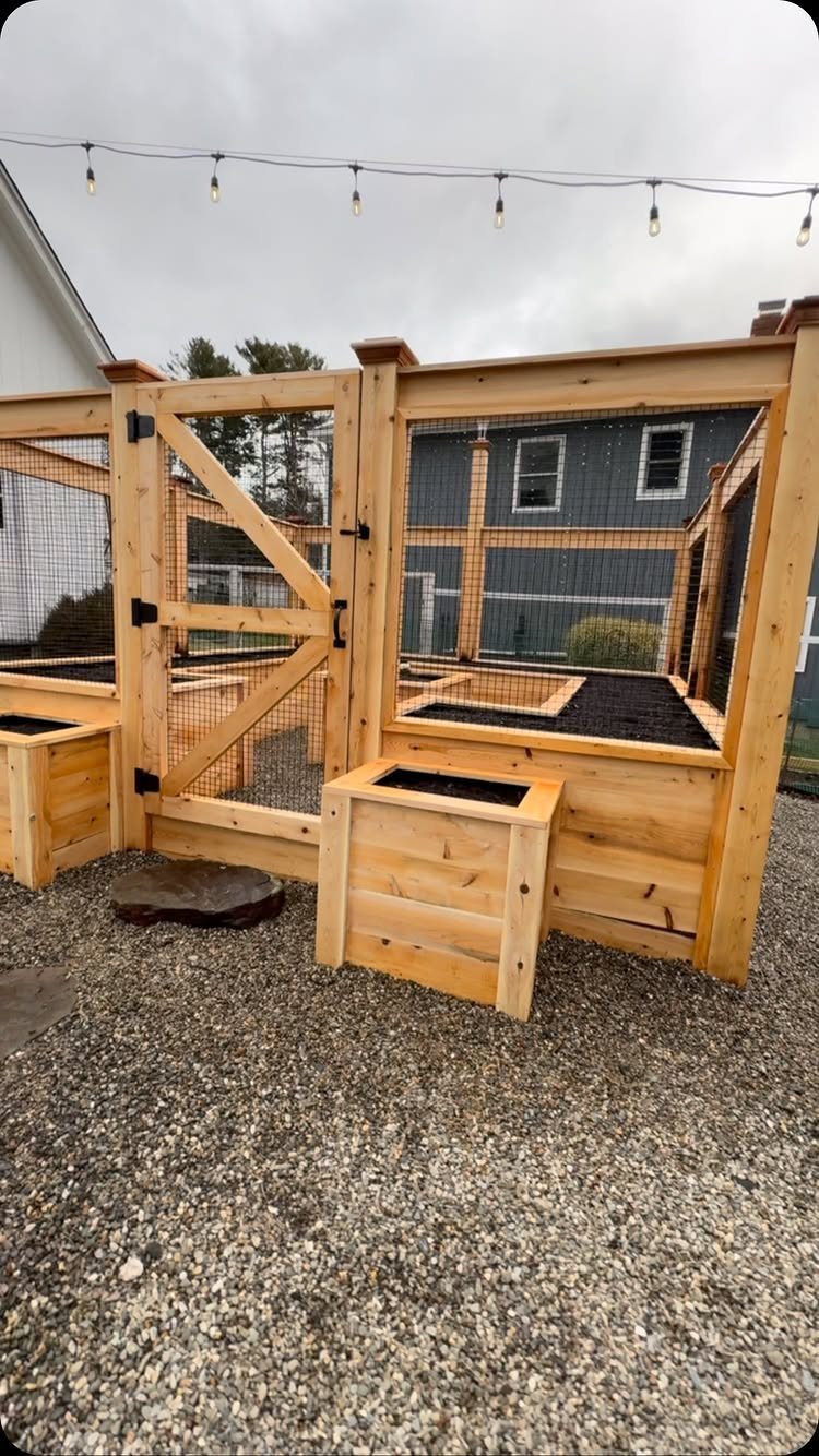 Wooden raised garden beds filled with soil sit inside a wooden fenced enclosure on gravel.