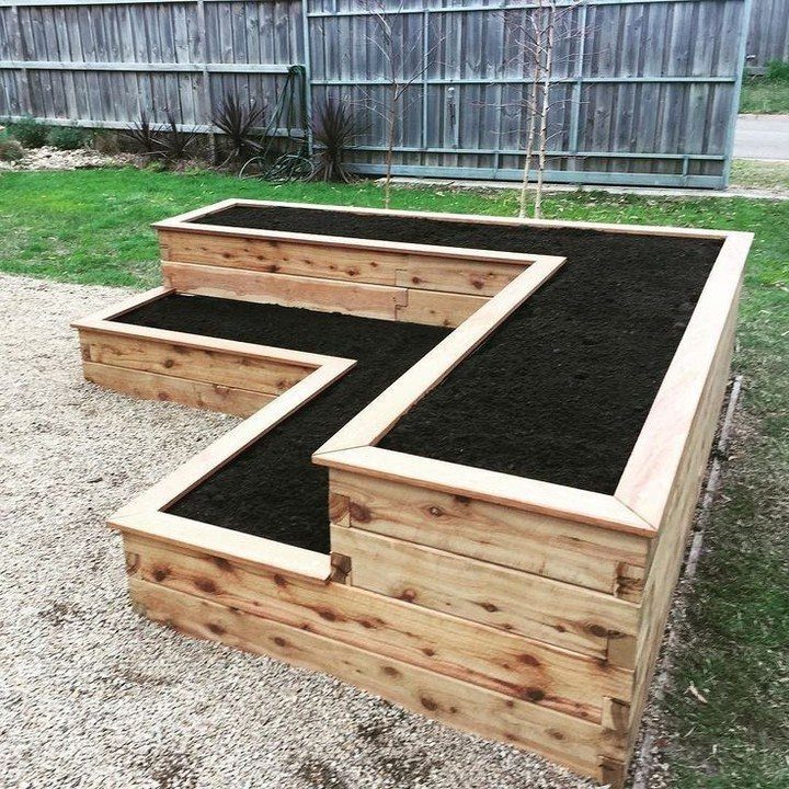 A large L-shaped wooden raised garden bed filled with dark soil on a gravel surface.