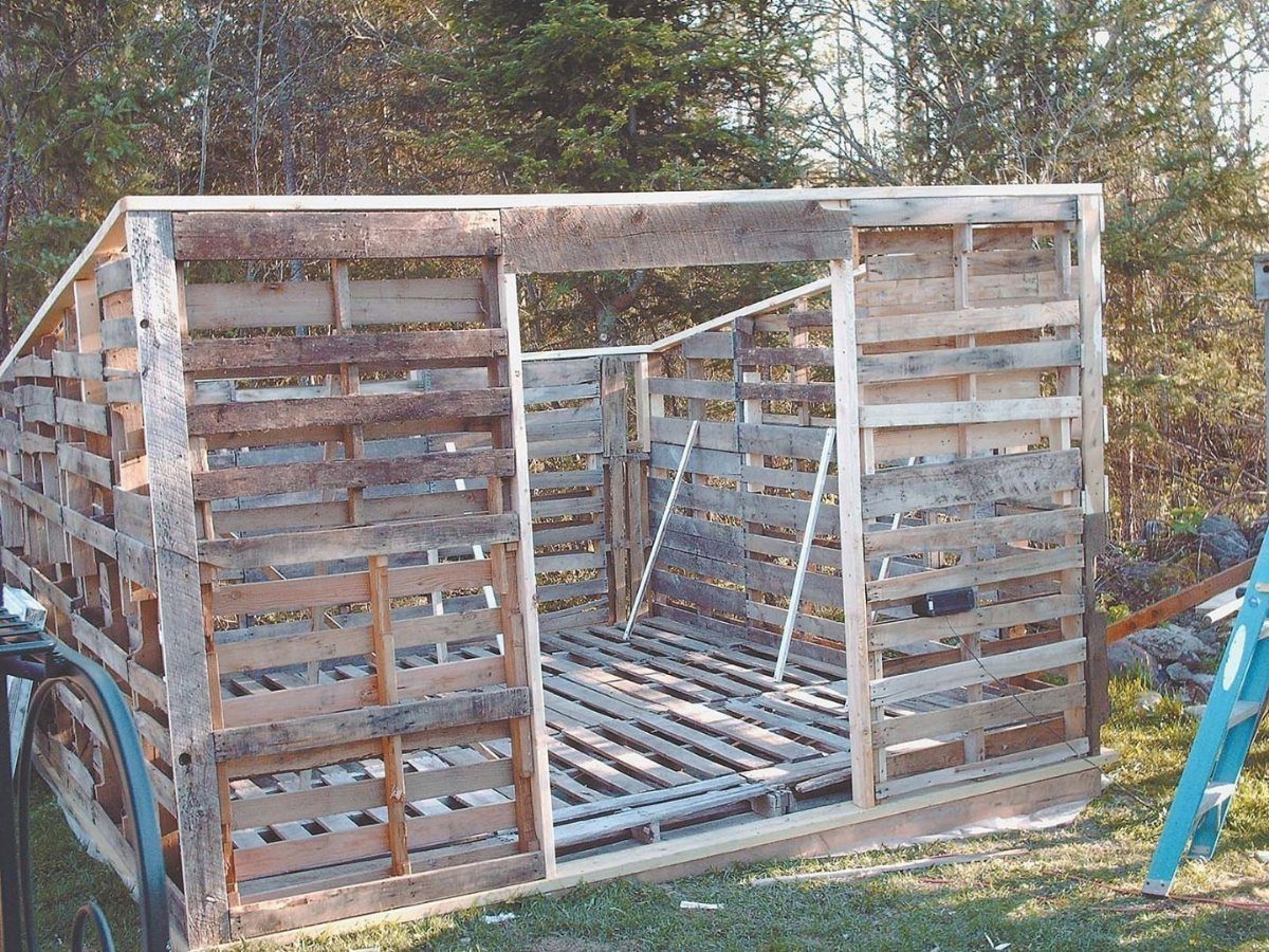 Greenhouse Using Pallets: 5 Builds for Small Yards 4 A rectangular structure constructed from wooden pallets sitting on a grassy area near trees.