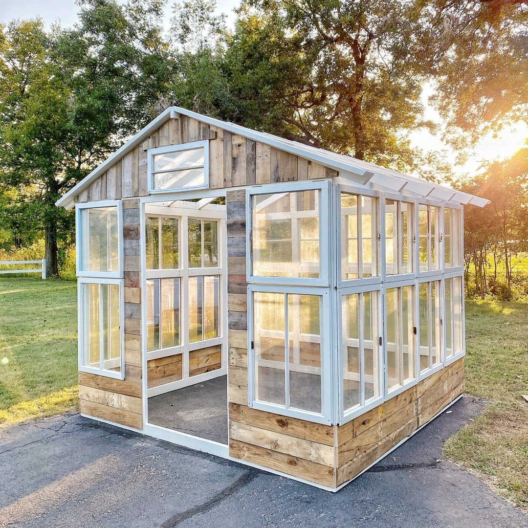 Greenhouse Using Pallets: 5 Builds for Small Yards 3 A small wooden greenhouse structure featuring many white-framed glass windows in a grassy outdoor setting.