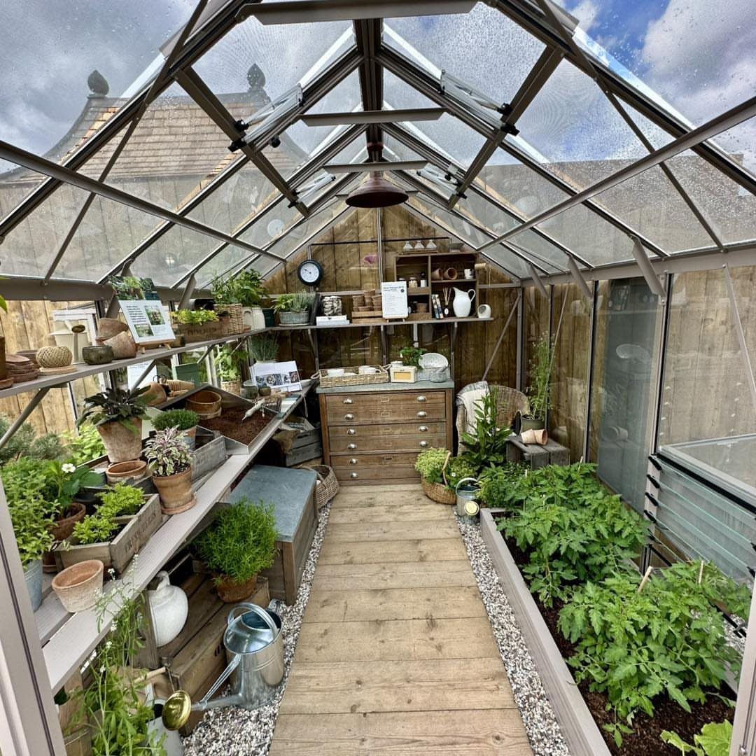 Interior view of a glass greenhouse filled with potted plants, wooden shelves, and a central walkway.