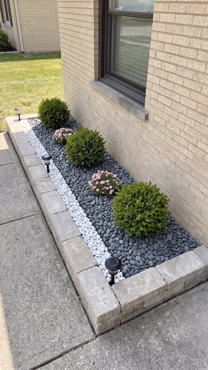 Dark grey pebbles and white pebble borders surround green shrubs and pink flowers in a stone garden bed.