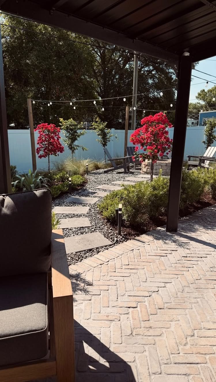 Herringbone brick pavers meet a gravel path with stepping stones and red flowering trees in a backyard.