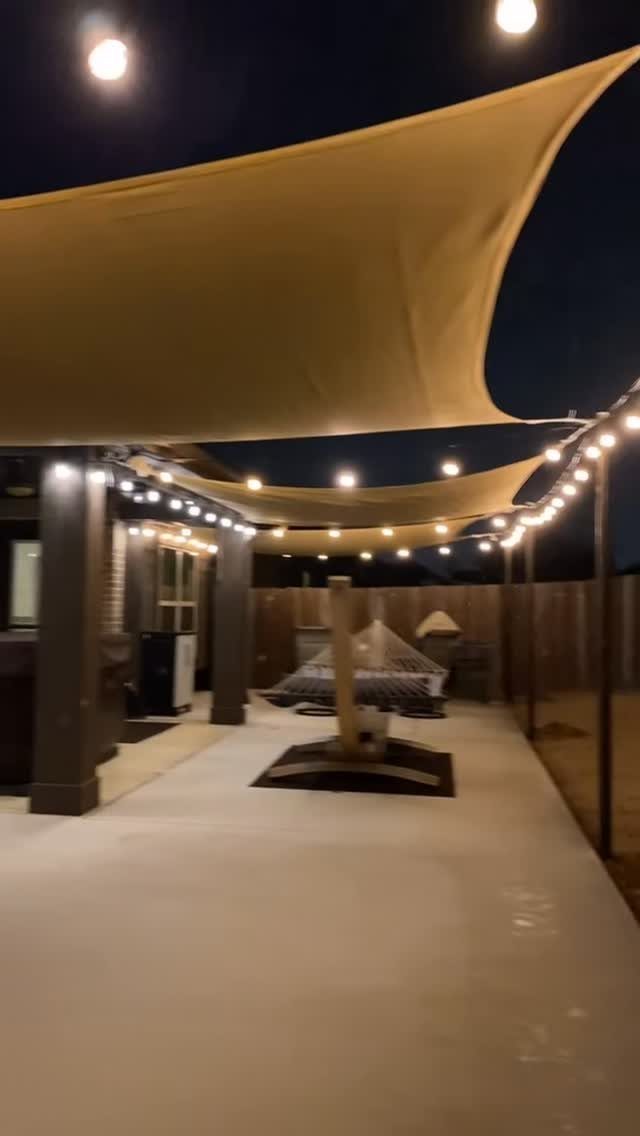 Tan shade sails and string lights illuminate a modern backyard patio at night.