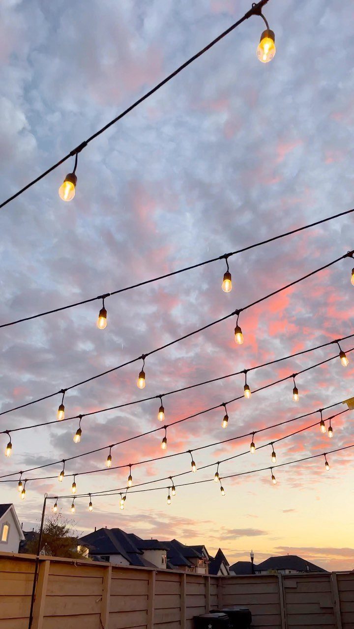String lights with glowing warm bulbs hang across a backyard under a pink sunset sky.