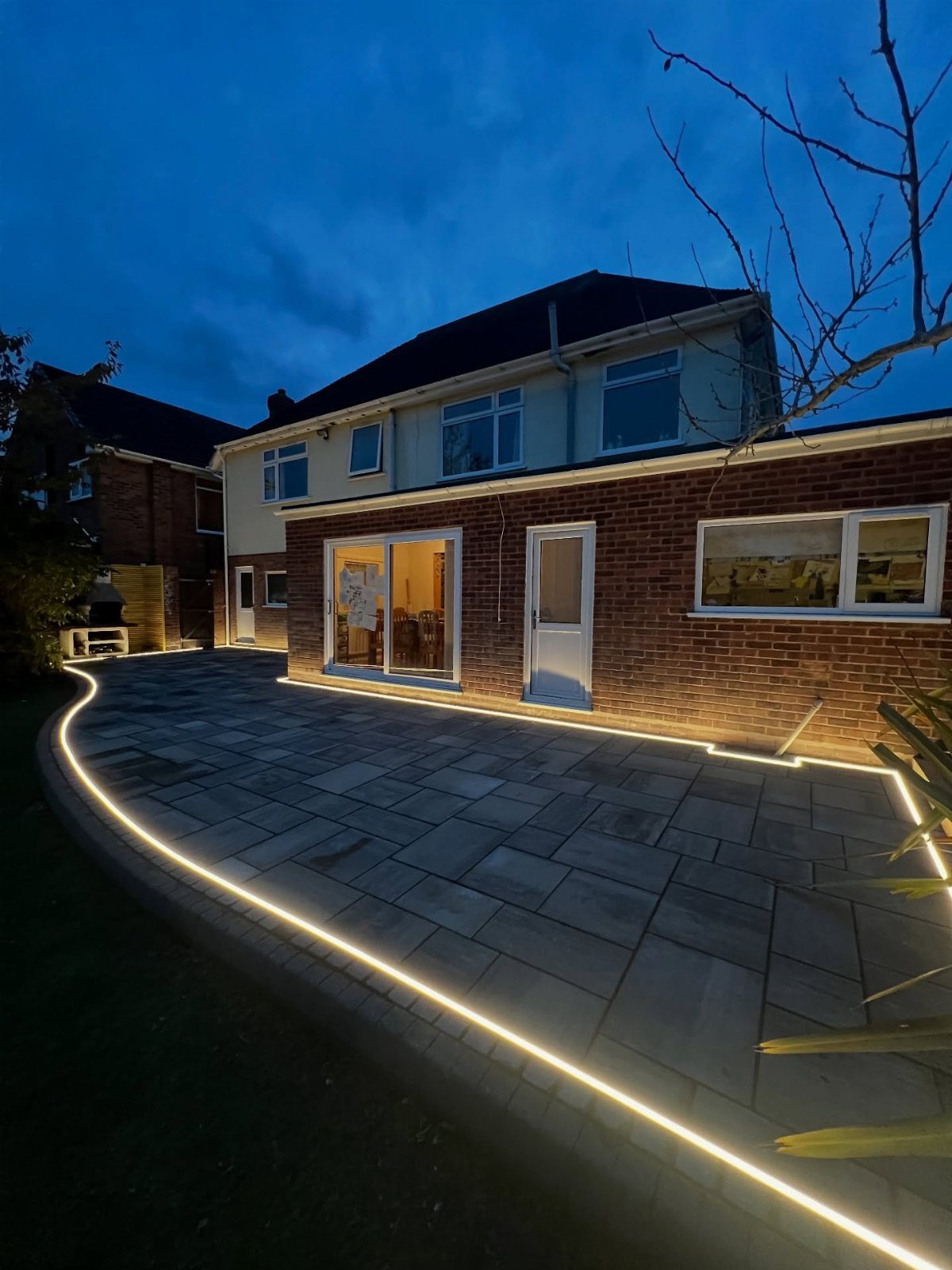 A large grey stone patio illuminated by glowing LED strip lights at dusk next to a brick house.