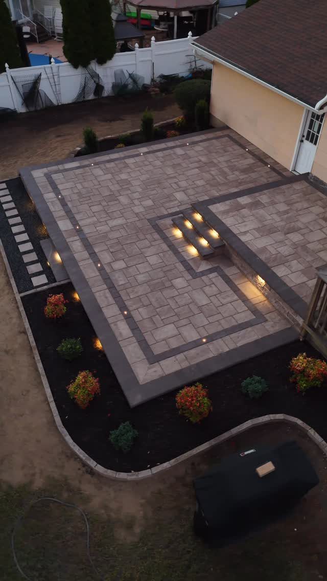 Large stone patio with grey pavers, dark borders, and integrated lighting next to a house.