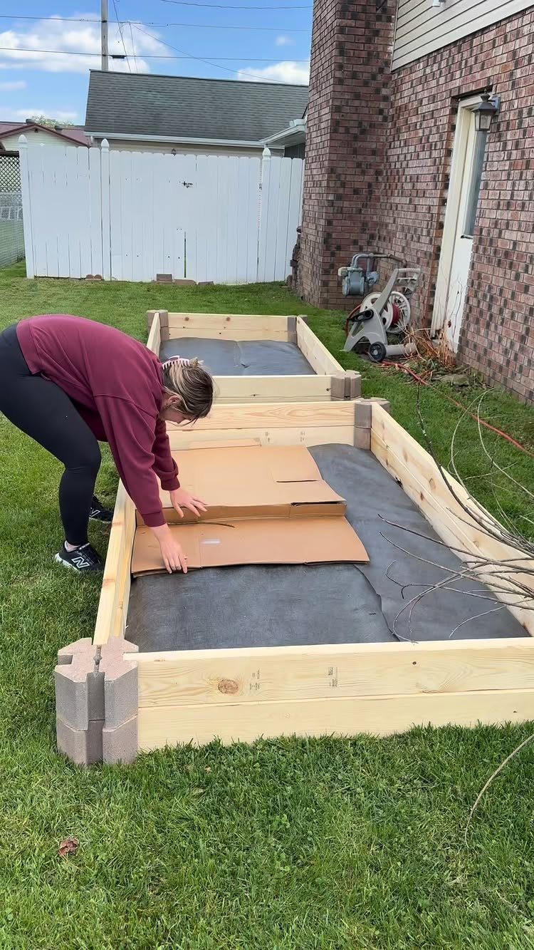 A person placing cardboard into a new wooden raised garden bed on a lawn.