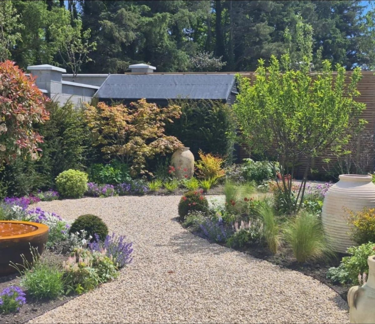 A gravel garden path winding through layered plants, shrubs, and tall trees.