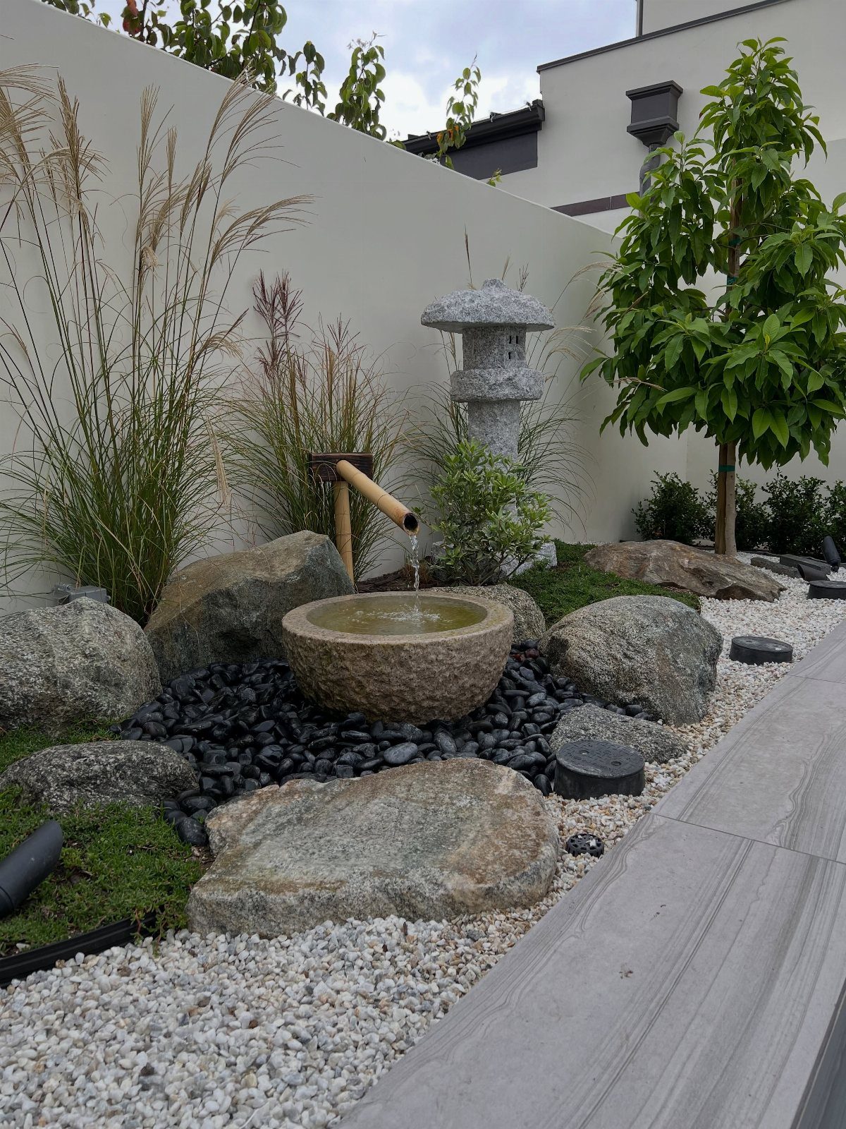 A stone water fountain with a bamboo spout pouring water into a basin surrounded by rocks.