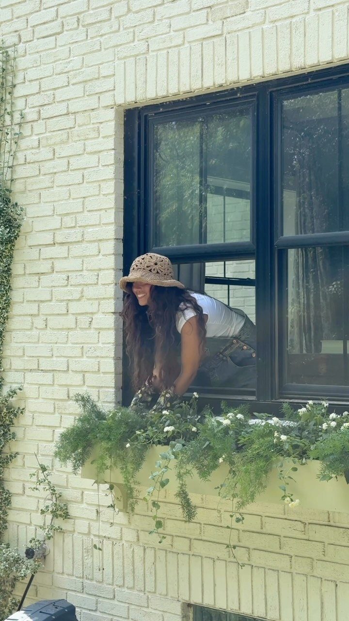 A woman smiling behind a window with a lush green window box below it.