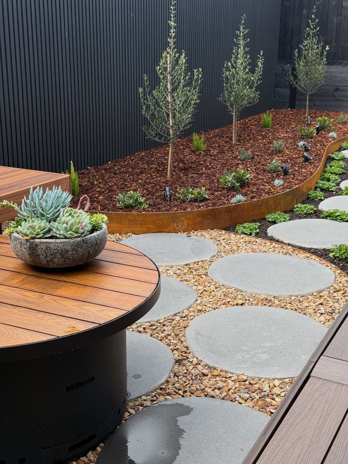 16 Small Home Landscaping Ideas That Fit Any Plot 14 Circular concrete stepping stones set in gravel leading to a landscaped garden bed with mulch and small trees.
