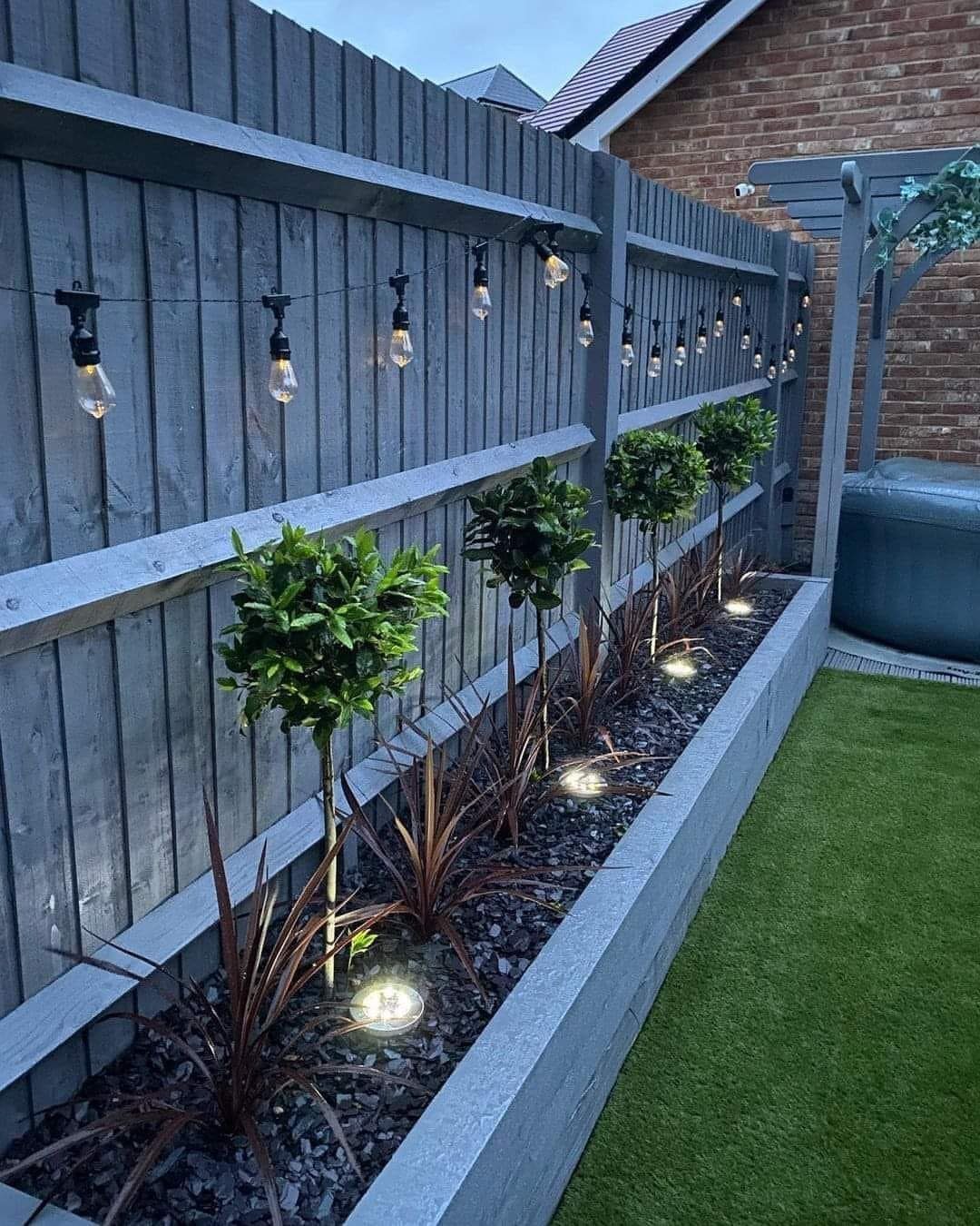16 Small Home Landscaping Ideas That Fit Any Plot 10 String lights hanging along a grey wooden fence above a landscaped garden bed.